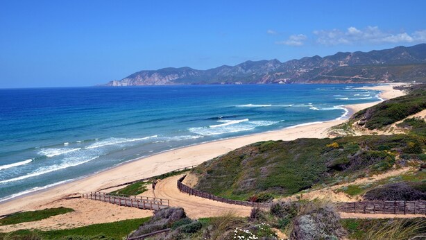 Un paisaje costero con playa de arena, una pasarela de madera y montañas al fondo.