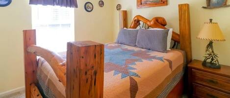 Una cama de madera con colcha, una mesita de noche con lámpara y una ventana con cortinas.