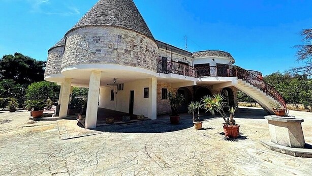 Ein Steinhaus mit einem Kegeldach, einem Balkon und einer Treppe, die zu einer oberen Ebene führt.