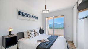Quarto com cama, mesa de cabeceira com abajur, janela grande e vista para o exterior.