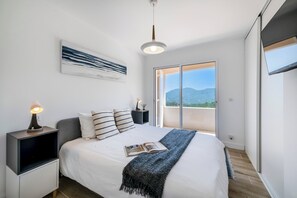 Ein Schlafzimmer mit einem Bett, einem Nachttisch mit Lampe, einem großen Fenster und Blick ins Freie.