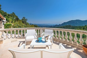 Eine Terrasse mit weißen Stühlen und einem Tisch, Blick auf die Landschaft, strahlend blauer Himmel.