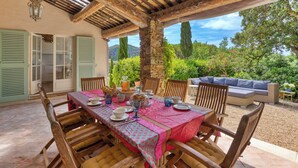 A covered outdoor dining area with a table set for a meal, wooden chairs, and a seating area with cushions. - CASA-Fontaine Luxury Villa in Gigaro (Cavalaire-sur-Mer)