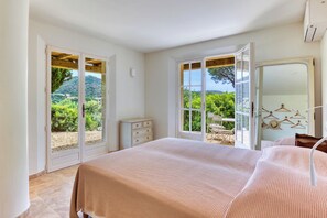 Ein Schlafzimmer mit einem Bett, einer Kommode und Blick auf Grün hinter offenen Türen.