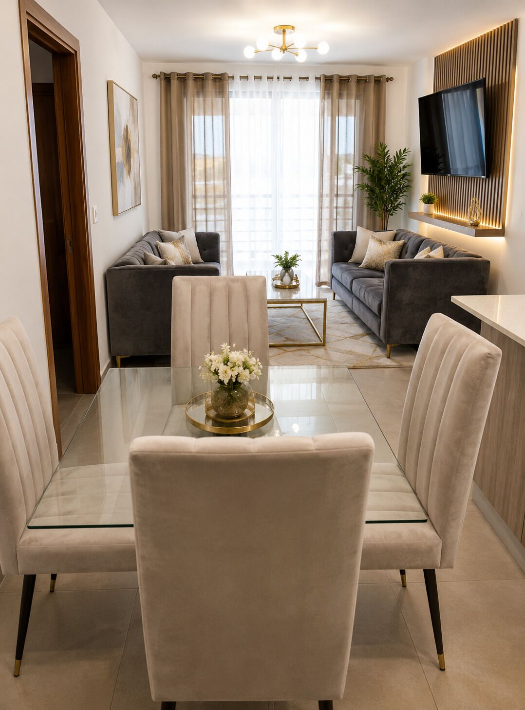 A modern living room with a glass dining table, two sofas, a TV mounted on the wall, and a potted plant.
