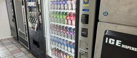 Uma fileira de máquinas automáticas com diversas bebidas, incluindo Pepsi e outros refrigerantes, e uma placa indicando 'DISTRIBUIDOR DE GELO'.