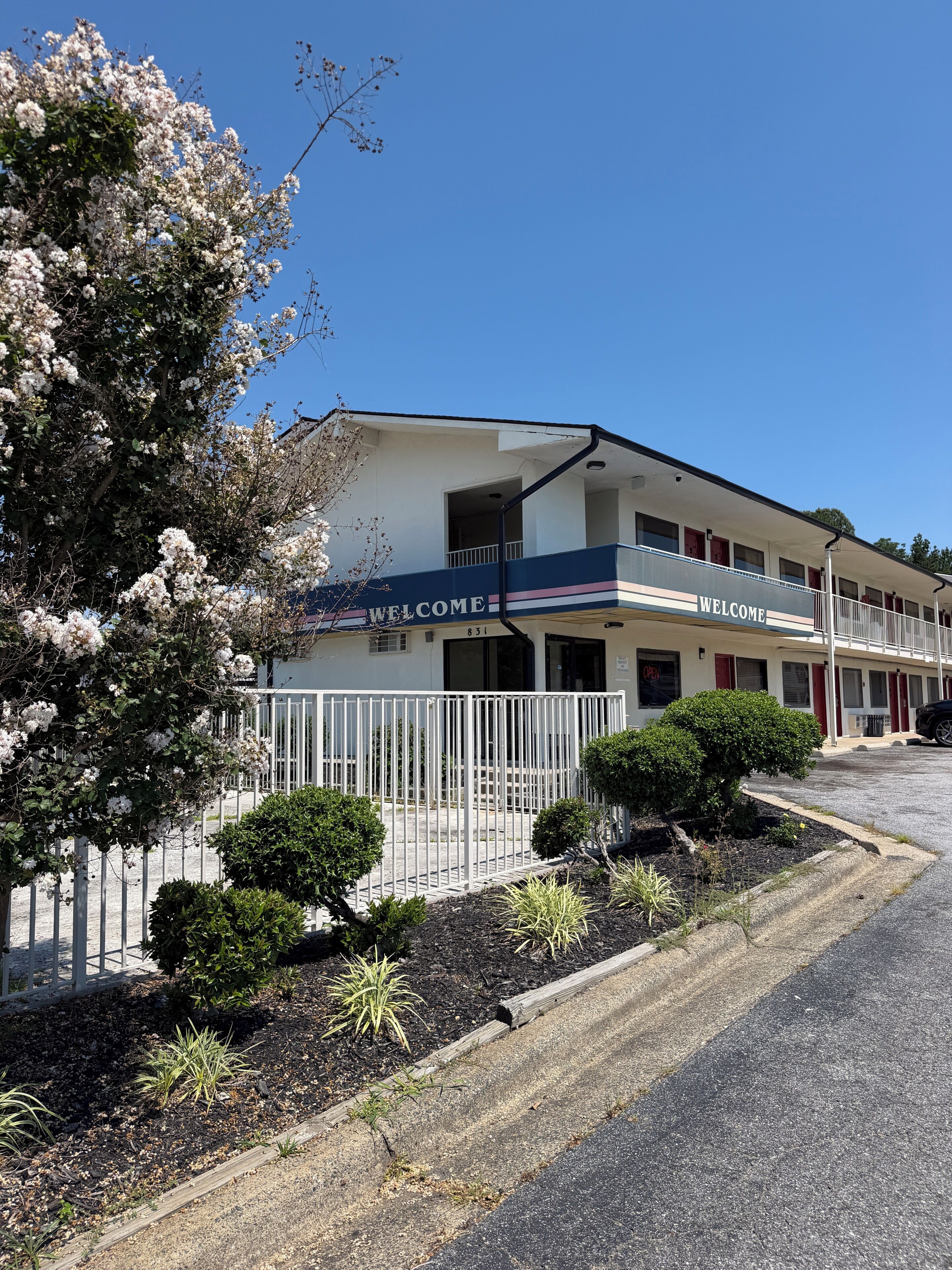 A motel with a 'WELCOME' sign, a fenced parking area, and a well-maintained garden.