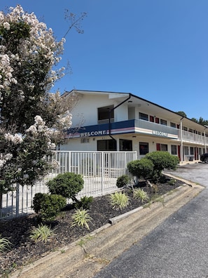 Un motel con un cartel de 'WELCOME', un estacionamiento cercado y un jardín bien cuidado.