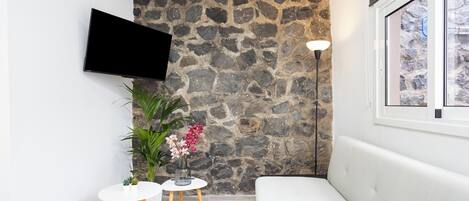 A modern living room with a stone wall, a flat-screen TV, a white sofa, a small round table with a vase of flowers, and a floor lamp.