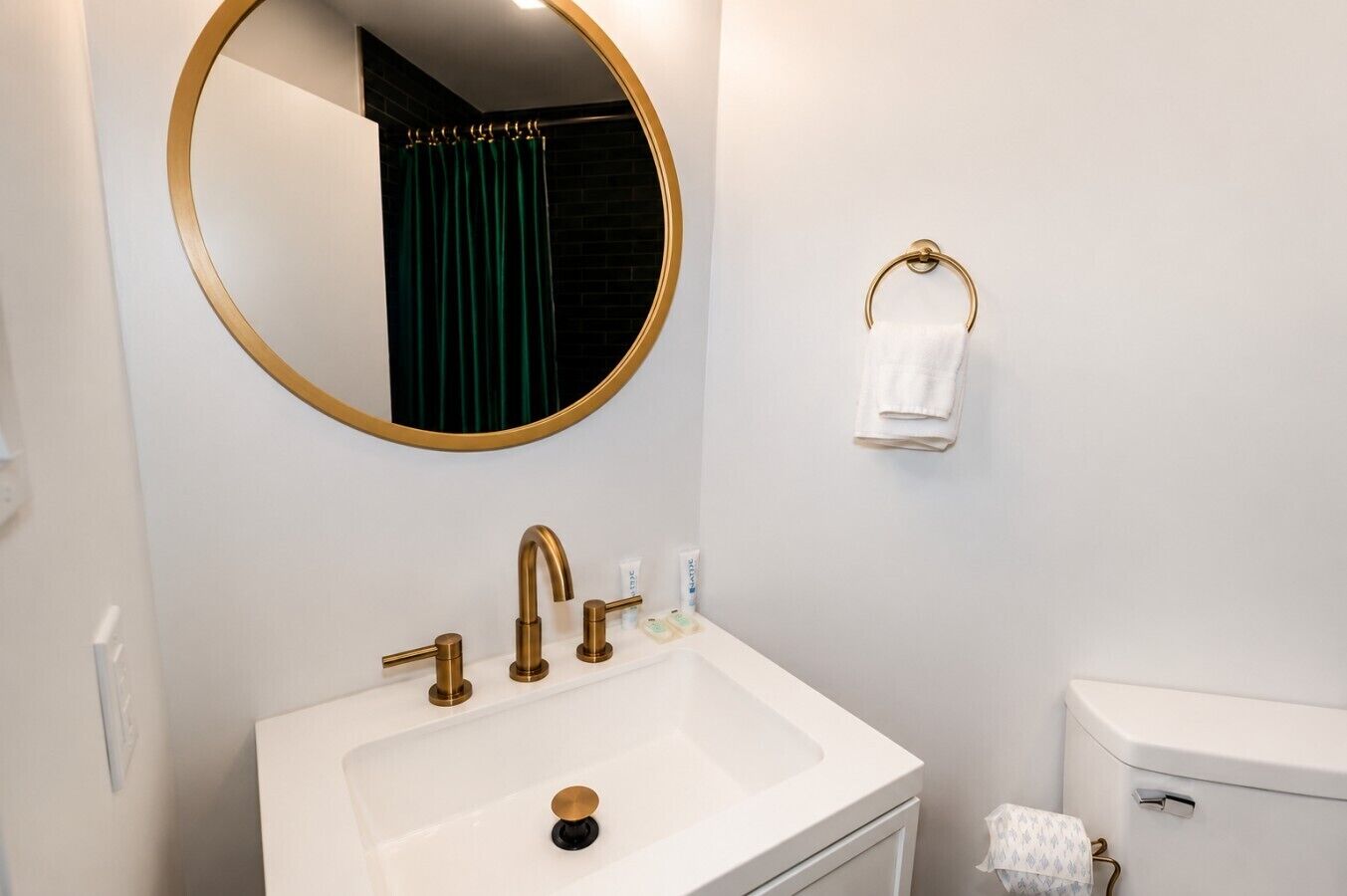 A bathroom with a white sink, gold faucets, a round mirror, and a toilet.