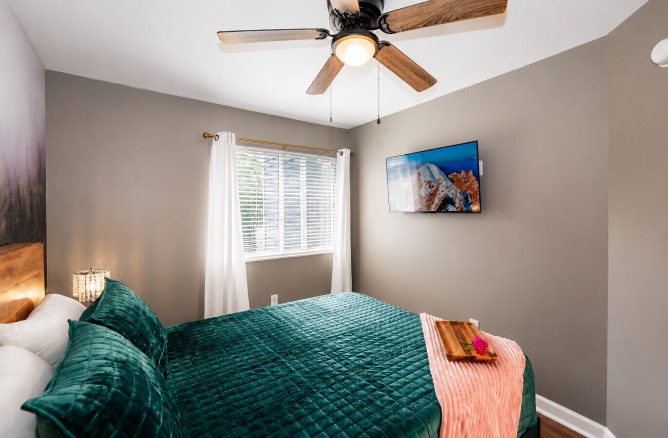 A bedroom with a bed, a ceiling fan, a window with blinds, a painting on the wall, and a bedside table with a lamp.