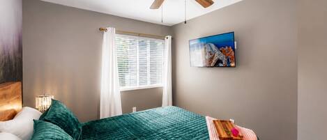 A bedroom with a bed, a ceiling fan, a window with blinds, a painting on the wall, and a bedside table with a lamp.