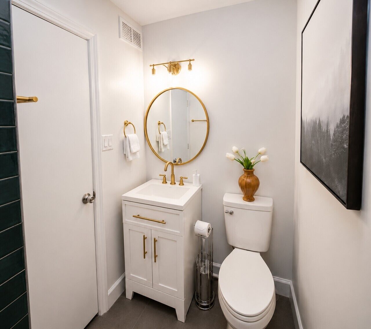 Ein Badezimmer mit einem weißen Waschbecken, goldenen Armaturen, einem runden Spiegel, einer Toilette und einem gerahmten Bild an der Wand.