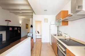 A modern kitchen with a built-in oven, a sink, and a stove. There is a small dining area with a chair and a table. The kitchen has wooden cabinets and a white countertop. - 23NO1138 - Apartment in Poble Sec (Barcelona)
