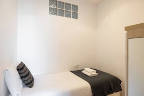 A small, single-bed room with a plain white wall, a window with frosted glass, and a wooden door. - 23NO1138 - Apartment in Poble Sec (Barcelona)