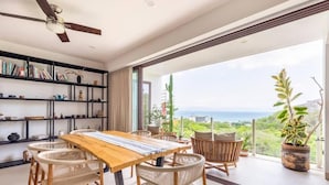 Ein moderner Essbereich mit einem Holztisch und Korbstühlen, einem Bücherregal mit verschiedenen Gegenständen und einem großen Fenster mit Blick auf das Meer.