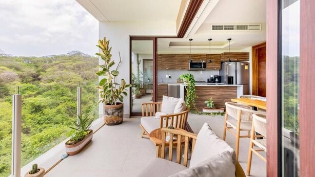 Ein Balkon mit Blick auf eine üppige Landschaft, Holzmöbel und eine moderne Küche, die durch eine offene Tür sichtbar ist.