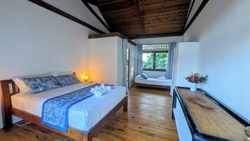 A bedroom with a wooden bed, a bedside lamp, a window with a view of greenery, and a small table with a vase of flowers.