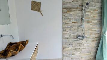 A bathroom with a shower, a wooden basin, a woven mat, and a stone wall.