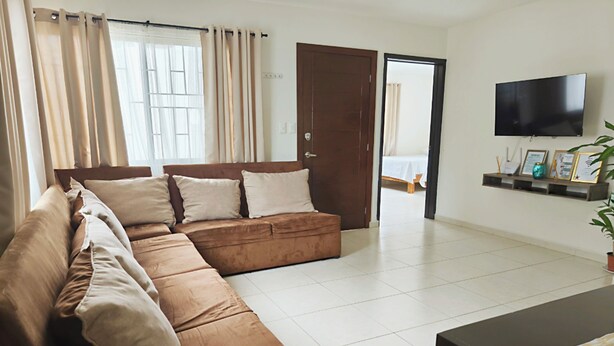 A living room with a brown sectional sofa, a flat-screen TV mounted on the wall, and a door leading to another room.