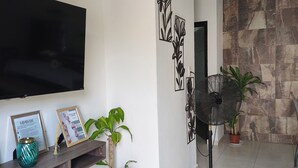 A living room with a flat-screen TV, decorative wall art, a potted plant, a standing fan, and a tiled floor.