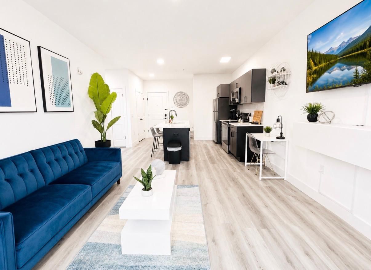A modern living room with a blue sofa, a white coffee table, and a kitchen area with dark cabinets and a white countertop.