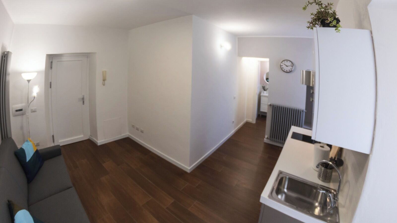 A modern living room with a grey sofa, wooden flooring, a kitchen area with a sink and countertop, and a white door.