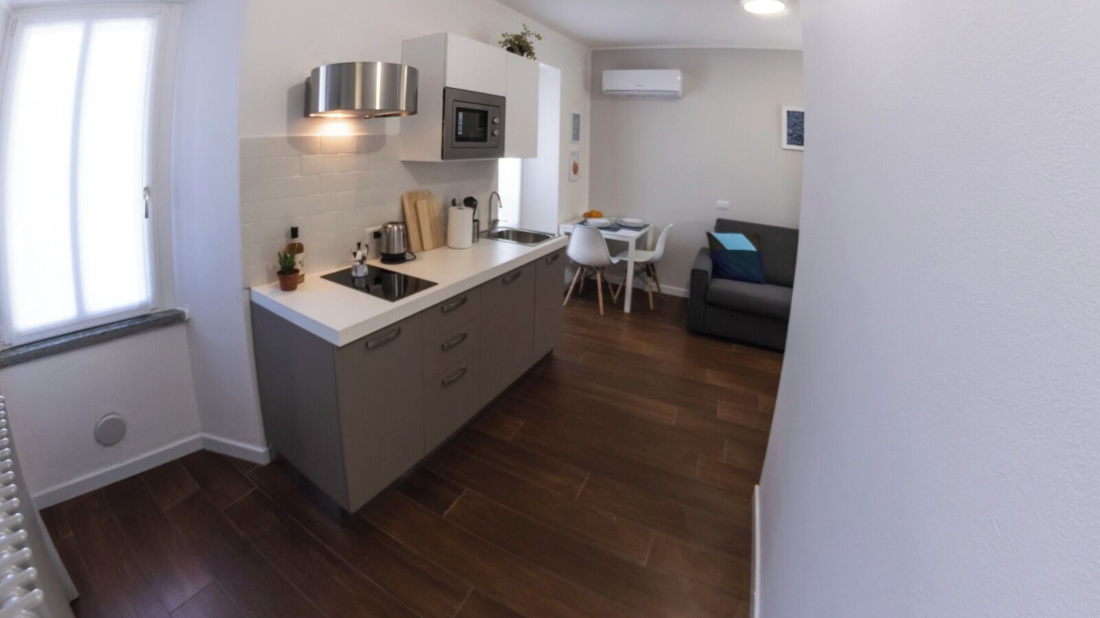 A compact kitchen with a microwave, sink, and cabinets, a dining area with a table and chairs, and a grey sofa.