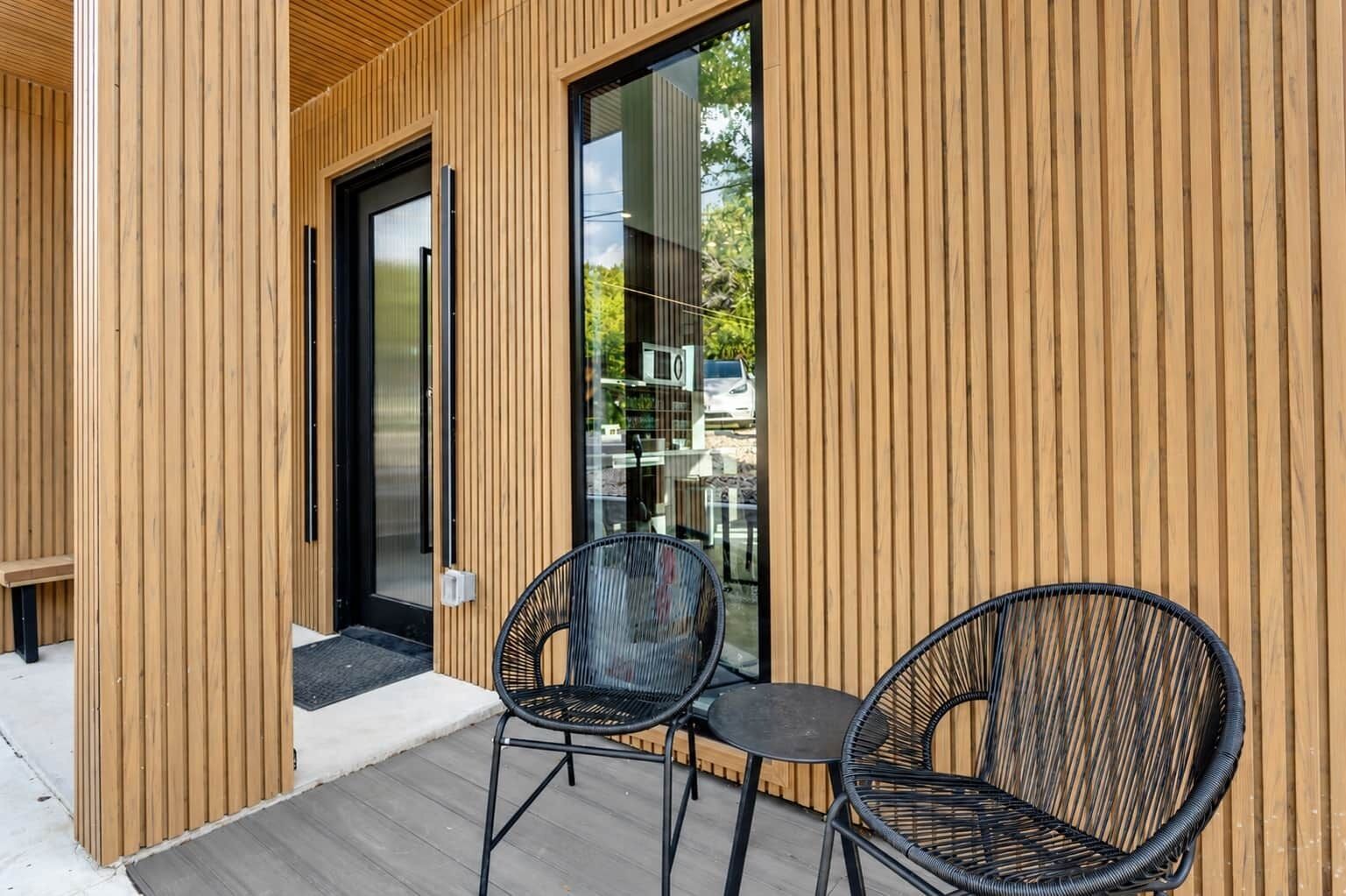 Une façade moderne avec un revêtement de bois vertical, une entrée avec une porte en verre et deux fauteuils en osier sur une terrasse en bois.