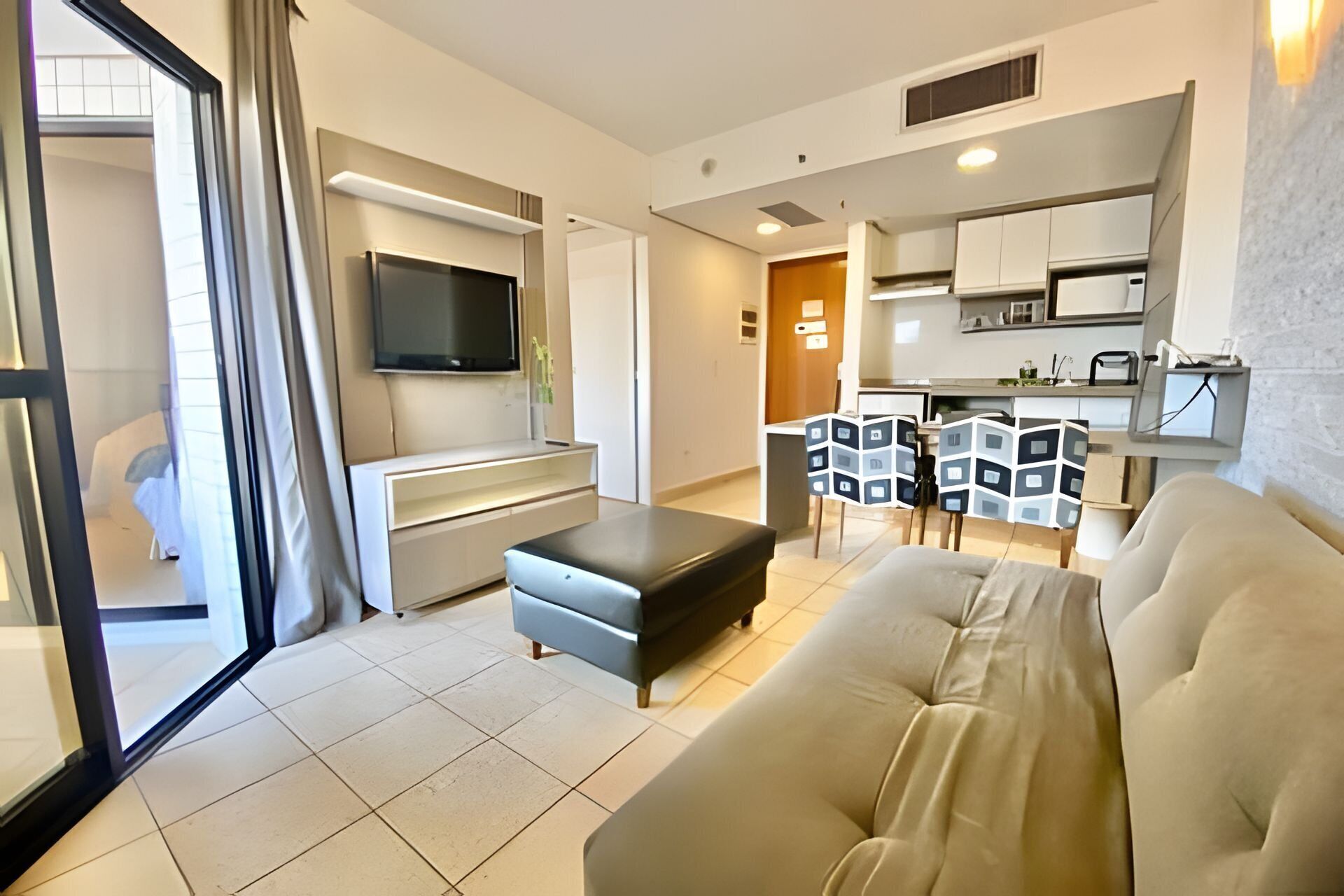 Een moderne woonkamer met een bank, een aan de muur bevestigde televisie, een eethoek met stoelen en een keuken op de achtergrond.