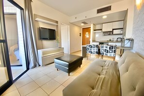 A modern living room with a sofa, a TV mounted on a wall, a dining area with chairs, and a kitchen in the background. - SHB Lindo apartamento frente a praia (Santos)