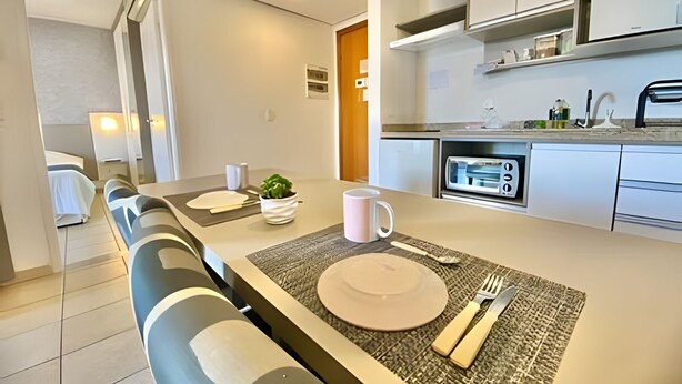 A modern kitchen with white cabinets, a built-in oven, and a dining area with a table set for two. - SHB Lindo apartamento frente a praia (Santos)