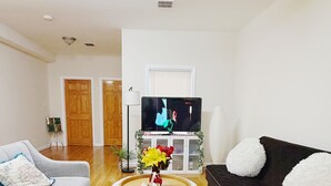 A living room with a grey sofa, a black sofa with white cushions, a white coffee table with a vase of flowers, and a flat-screen TV on a stand. - Beautiful Private room in a  2-bedroom apartment in vibrant Jersey City (Jersey City)