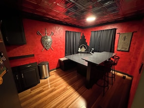 A room with a red wall, a wooden floor, a table with chairs, a trash bin, and a ceiling with a grid pattern.
