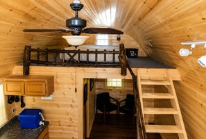 L’intérieur d’une cabane comprend un espace nuit en mezzanine, un ventilateur de plafond, une fenêtre, un coin cuisine avec des placards et une glacière bleue.
