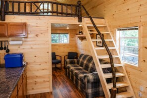 L’intérieur d’une cabane avec un escalier en bois, un canapé à carreaux, un coin cuisine et une fenêtre donnant sur l’extérieur.