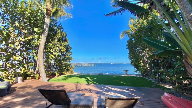A deck with two wicker chairs, a small table, and a view of the ocean with a pier in the distance. - Waterfront cottage in Jensen Beach perfect for fishing, kayaking, and relaxing! (Jensen Beach)