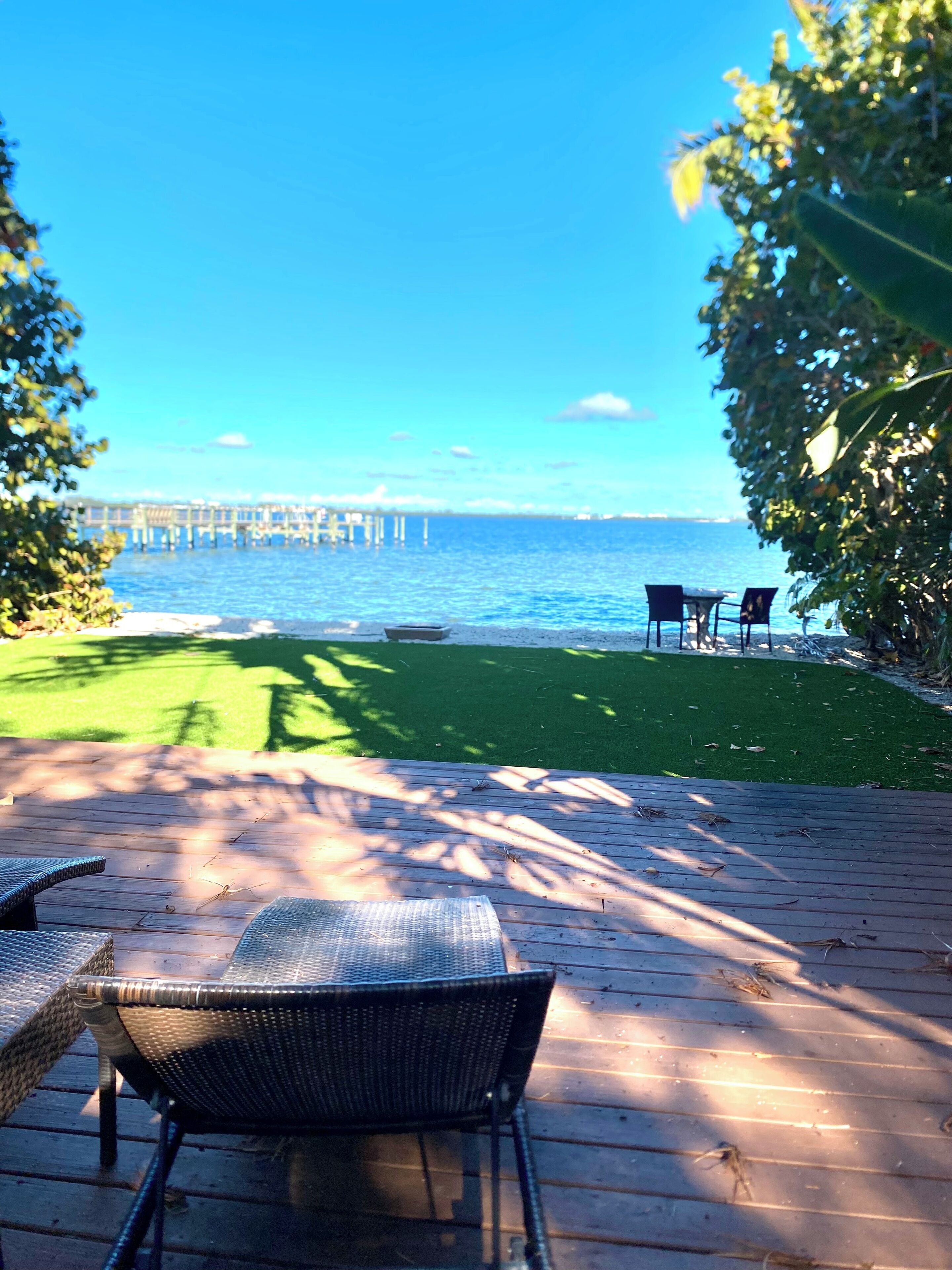 A deck with chairs and a table, overlooking a grassy area and a pier extending into the water.