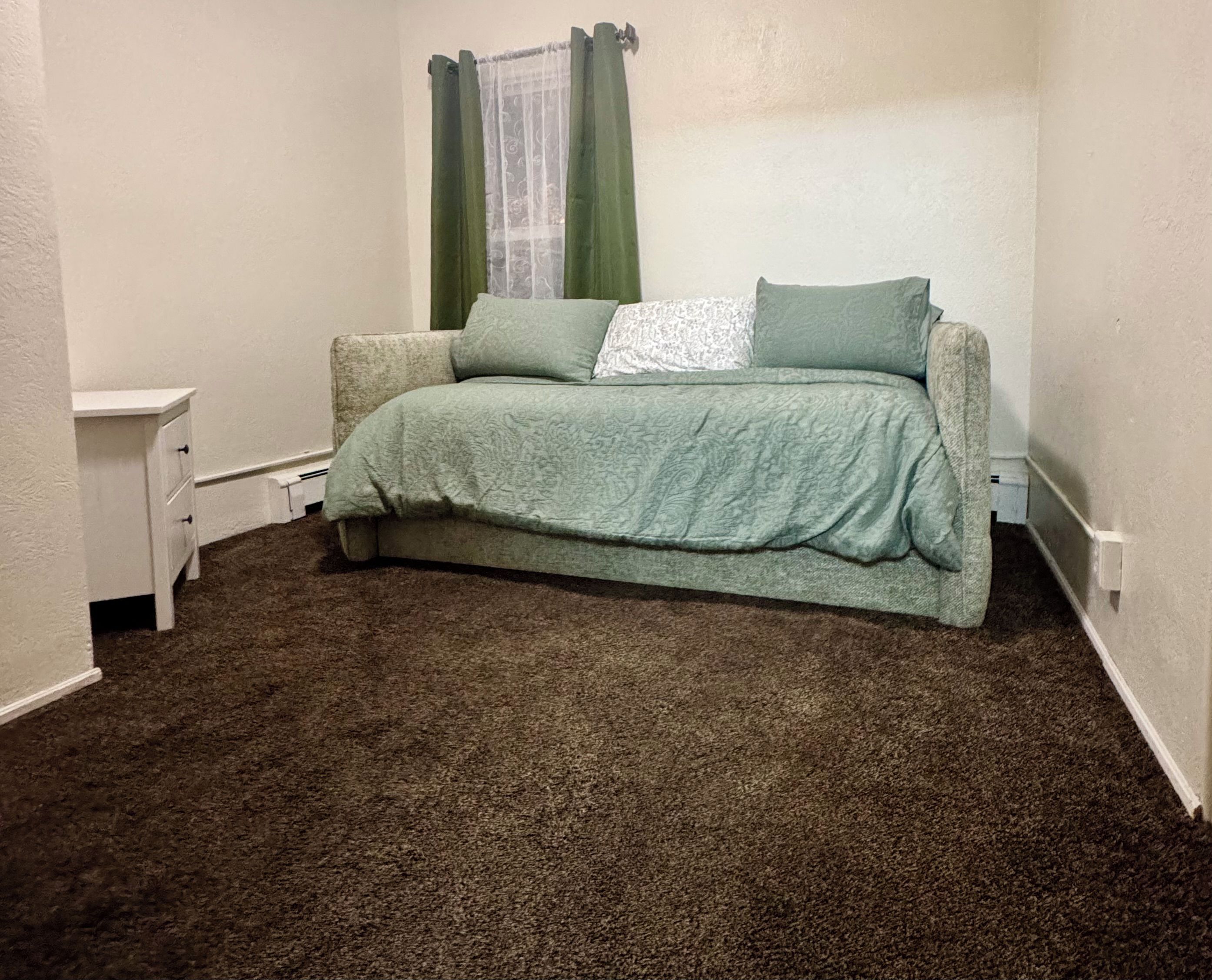A small, single-bed room with a green bedspread, a nightstand, and a window with green curtains.