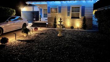 A residential driveway with a white car parked on the left, a stone fountain in the center, and a house with blue shutters and a stone facade in the background.
