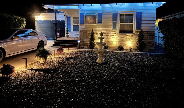A residential driveway with a white car parked on the left, a stone fountain in the center, and a house with blue shutters and a stone facade in the background.