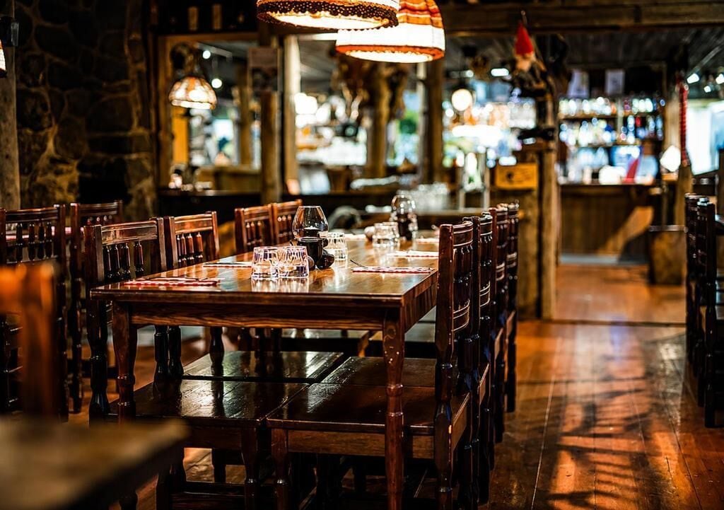Un restaurante con una larga mesa de madera y sillas, lámparas colgando del techo y una barra al fondo.
