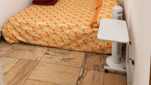 Une petite pièce, de taille modeste, avec un lit superposé, un lit simple avec une literie à motifs, un oreiller rouge, une petite table avec une lampe et un radiateur posé sur le sol.