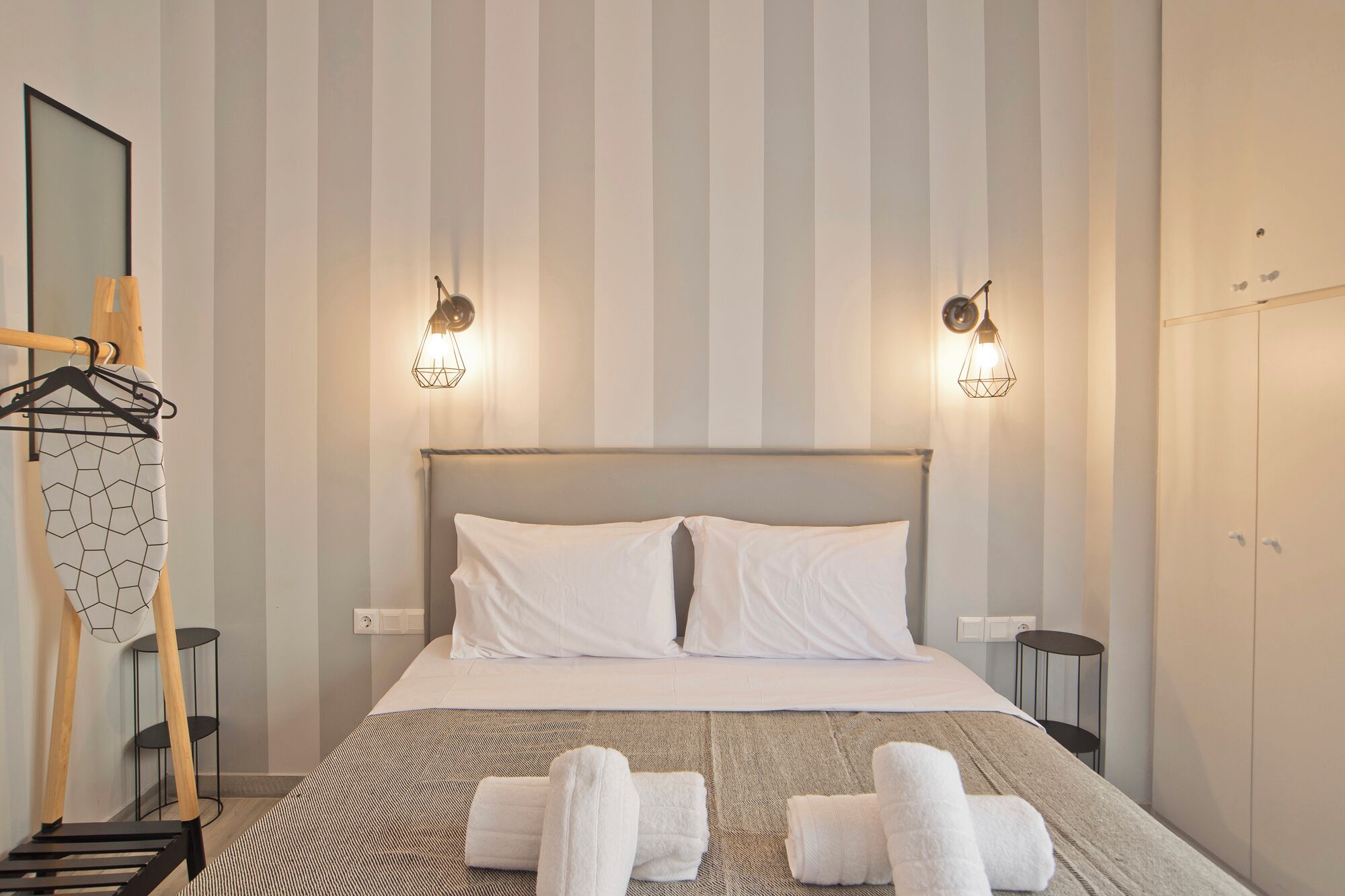 A hotel room with a striped wall, two bedside lamps, a bed with white linens, a wooden headboard, and a black metal hanger with a white towel.