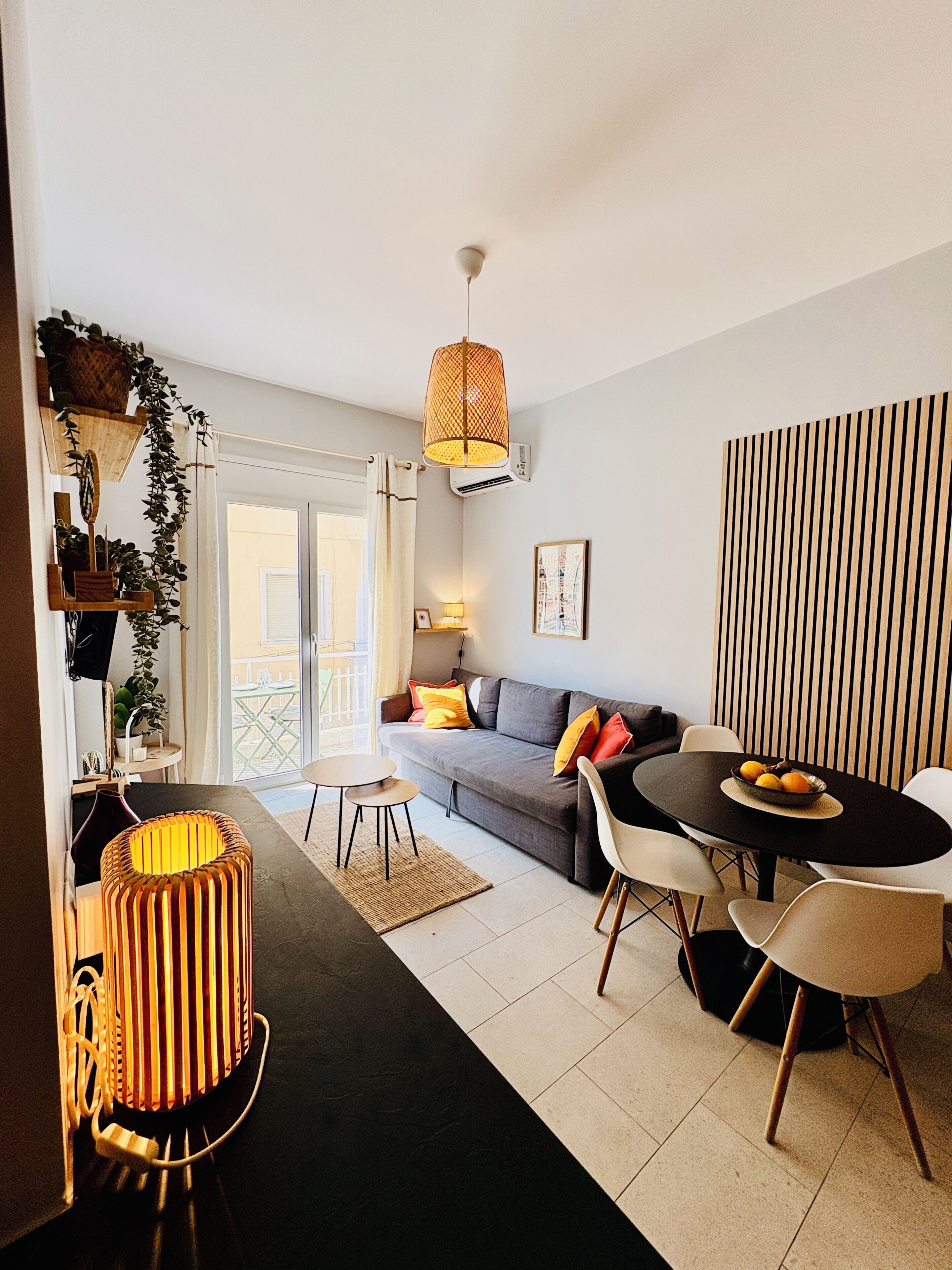 A modern living room with a sofa, dining table, and chairs. There is a hanging light fixture and a wall-mounted air conditioner.
