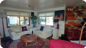 Un salon avec deux canapés blancs, une table basse ronde et un divan rouge. On y trouve des fenêtres avec des stores, une plante en pot et une lampe sur une table d’appoint.