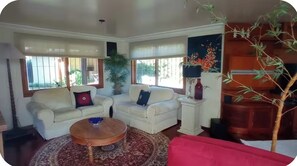 Ein Wohnzimmer mit zwei weißen Sofas, einem runden Couchtisch und einem roten Sitzsofa. Es gibt Fenster mit Jalousien, einen Topfpflanzen und eine Lampe auf einem Beistelltisch.