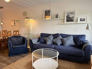 A living room with a blue sofa, a glass coffee table, and a dining area with wooden chairs and a table. - 5 Star Holiday Home in Uddevalla (Uddevalla)