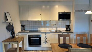 A compact kitchen with white cabinets, a built-in oven, a microwave, and a coffee maker. There is a wooden table with two chairs and a small wooden shelf with a wine bottle and glasses. - 5 Star Holiday Home in Uddevalla (Uddevalla)