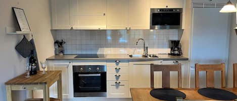 A compact kitchen with white cabinets, a built-in oven, a microwave, and a coffee maker. There is a wooden table with two chairs and a small wooden shelf with a wine bottle and glasses.